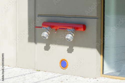 Red fire hydrant with two silver outlets mounted on a gray wall, accompanied by a warning sign and circular markers on the ground near a glass building
