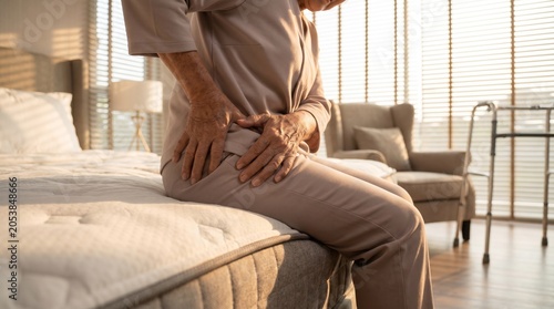 Elderly woman sitting on bed feeling chronic hip pain at home in bedroom