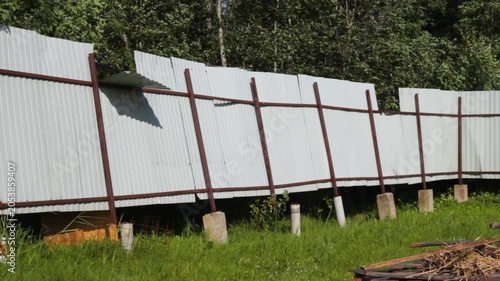 Still frame of a damaged corrugated metal fence on a rural property. Loose and bent panels sway and rattle in gusty wind while nearby trees and vegetation move in the background