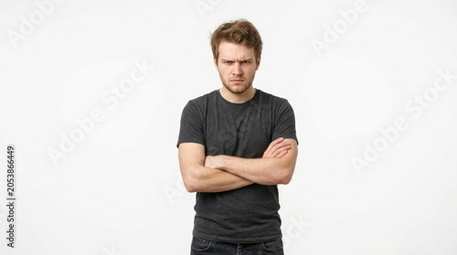 A young man with tousled brown hair stands with his arms crossed, displaying a displeased expression. He is wearing a gray t-shirt and dark jeans.