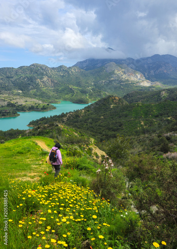 Woman with backpack hiking a trail overlooking a reservoir dam nestled among green mountains with spring wildflowers