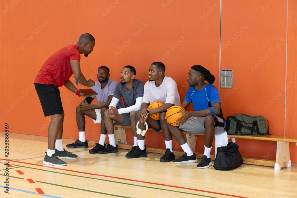 Fototapeta premium Diverse male team in sportswear sitting on bench in orange gym coach pointing tablet holding balls