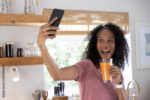 African American woman holding smartphone sipping orange juice and taking selfie at kitchen window