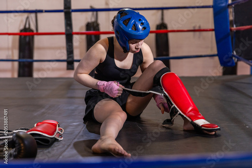 Red shin guard being fastened on padded boxing ring floor with blue headgear nearby