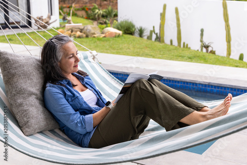 Senior woman reclining in striped hammock reading paperback on pool deck wearing denim shirt watch