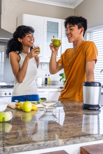Diverse couple savoring green smoothies after blending, clinking glasses and laughing in kitchen