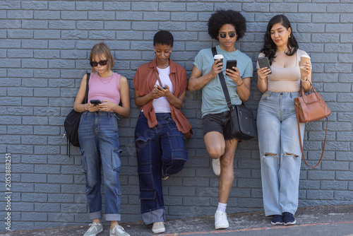 Diverse friends leaning on bluish brick wall on sidewalk, checking smartphones and holding coffee