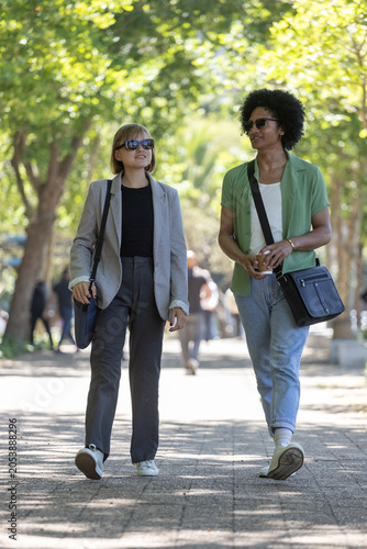 Two adults walking and talking on dappled tree-lined path, carrying crossbody bags and coffee cup