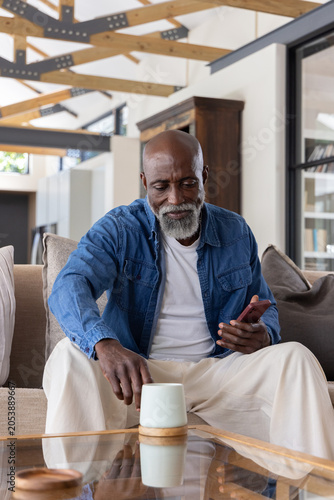 Senior African American man sitting at home in denim shirt holding smartphone and ceramic cup