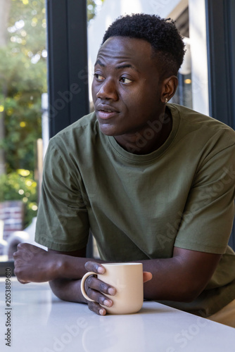 African American man sitting at countertop in olive tee holding beige mug gazing left toward glass
