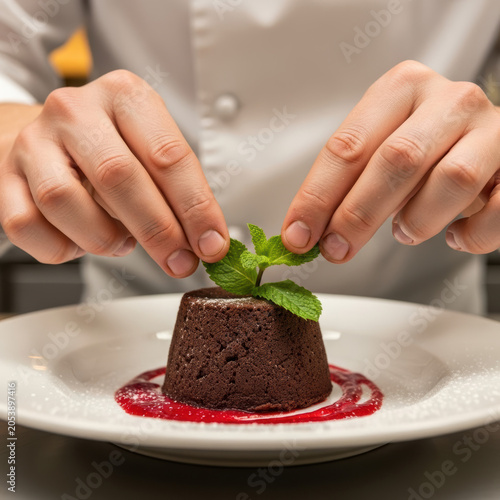 Professional chef hands placing fresh mint leaves on a delicious chocolate lava cake dessert on a white plate in kitchen