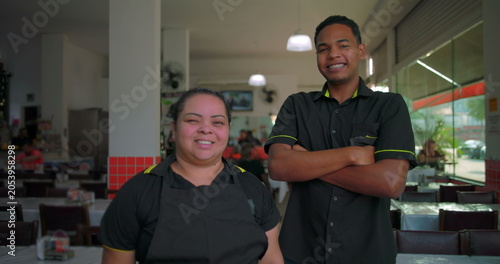 Latin employees smile together inside restaurant dining area, standing in uniform during lighthearted break in casual midday shift at urban food service spot
