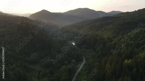 Aerial view cinematic shot of amazing mountains and warm sunset. Beautiful green forest slopes of mountain range. Bright disk of sun in sky slowly sets tops of mountains at summer. Nature concept
