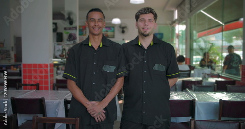 Two restaurant waiters smiling while standing inside dining area, hands folded and behind back, glass wall and tables visible in background
