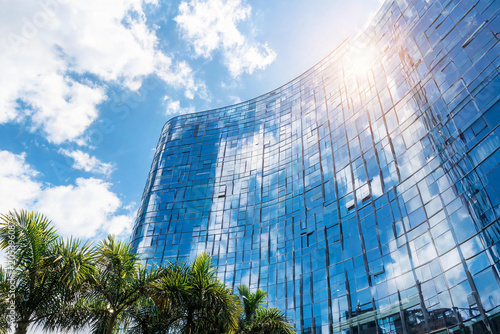 Modern Curved Glass Office Building Reflecting Blue Sky and Clouds with Palm Trees in Foreground