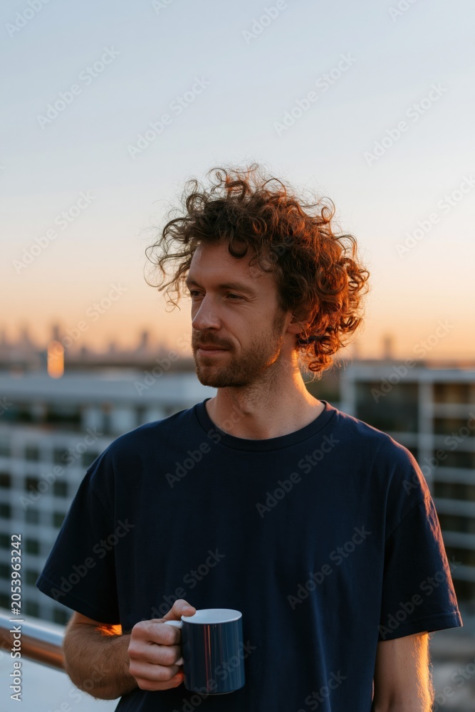 Naklejka premium Young man with curly hair holding a blue mug on a rooftop at sunset for lifestyle portrait and morning routine background for wellness and relaxation banner
