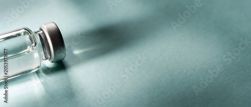 Glass vial with clear medicine solution lying on its side on a light teal surface casting a soft shadow.