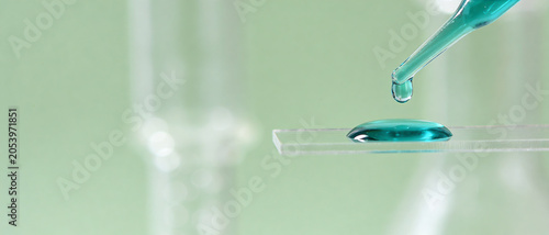 Glass pipette dripping teal liquid onto a laboratory glass slide against a green background.