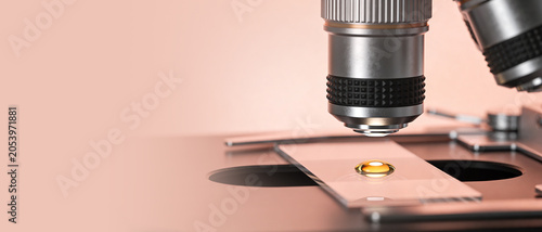 Close-up shot of a microscope objective over a glass slide with a glowing amber liquid droplet on a pink background.
