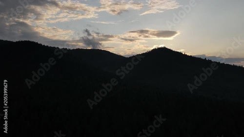 Aerial drone footage of silhouette amazing mountains and cloudy sky at sunset. Forest slopes of mountain range. Bright disk of sun slowly sets tops of mountains. Dramatic sunrise sky background