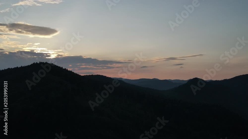 Aerial drone view flight over beautiful old spruce tree forest in mountains at sunset. Bright disk of sun slowly sets tops of mountains. Dramatic sunrise sky background