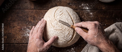 Photos Baker scores sourdough bread dough with lame, preparing loaf for baking, traditi