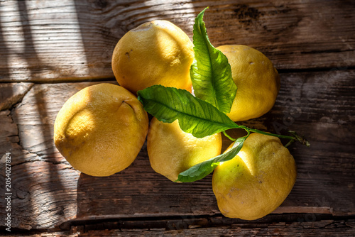 Limones amarillos enteros frescos con hojas verdes sobre mesa de madera rústica con luz solar directa y sombras. Toma en plano cenital con iluminación natural dramática de ventana y sombras marcadas.