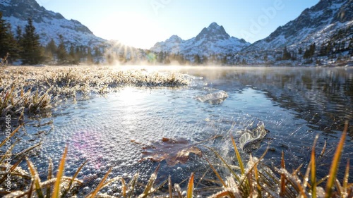 As the sun rises over the serene lake, golden rays illuminate the crystal-clear water, reflecting the towering mountains and enhancing the tranquil atmosphere of the serene landscape.