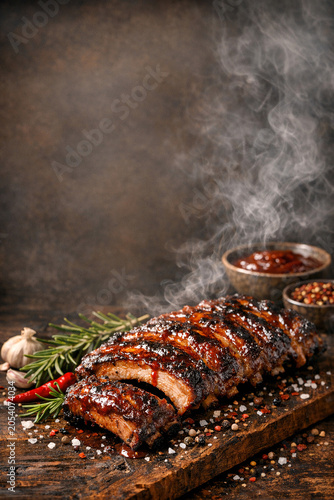 Smoky grilled ribs on wooden board. Steam rises from juicy meat. Garlic, rosemary, chili peppers nearby. Seasonings and spices scattered around. Rustic dark background enhances flavor