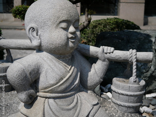 This image shows a gray stone sculpture of a smiling child monk carrying a bucket on a stick.