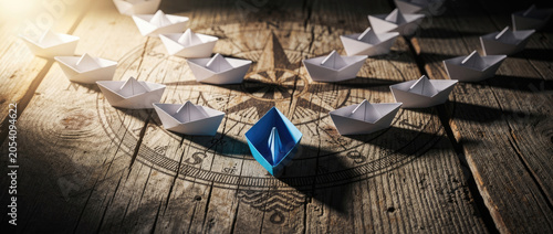 A single blue paper boat leads a fleet of white paper boats on a wooden surface.