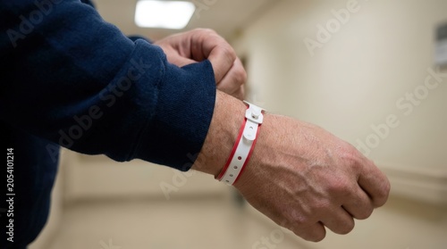 Close up shot of a person wearing a white and red plastic identification wristband on their arm while adjusting their dark blue sleeve inside a bright clinical hospital hallway setting.