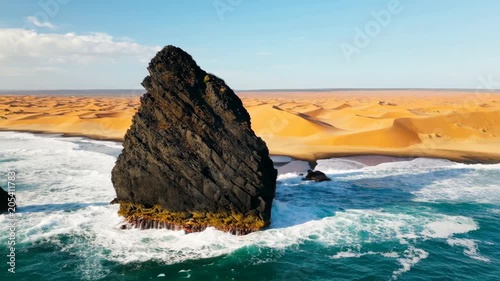Large black rock standing in the blue ocean next to golden sand dunes on a sunny day