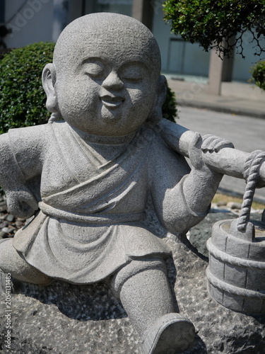 This image features a stone statue of a smiling child monk carrying a bucket on a stick, commonly found in outdoor temple gardens.