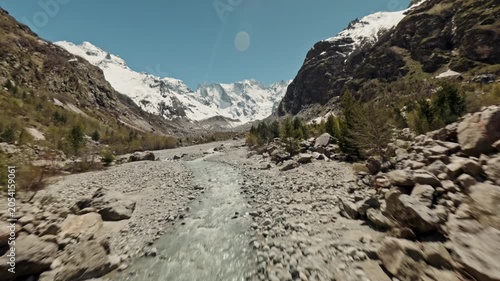 Fast-moving river racing through a rocky alpine valley toward the viewer, framed by large boulders and pale stones in the foreground, conifer-covered slopes