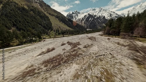 The Fpv drone flies low over the trees that grow abundantly along the mountain river. In the distance, the white peaks of the snow-capped mountains are visible. High quality 4k footage