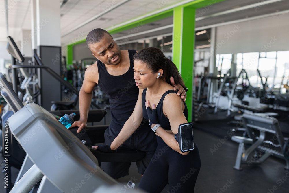 Fototapeta premium Young woman experiencing discomfort during a treadmill workout while supportive trainer assists her in modern gym, emphasizing fitness safety and exercise awareness