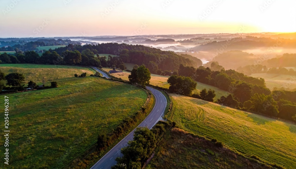 Fototapeta premium Scenic country road winding through rolling hills at sunrise, misty valley.