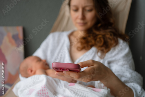 Mother cradling a newborn baby in one arm, actively using a smartphone with the other hand, depicting modern parenthood, multitasking, and digital connectivity in the postpartum journey