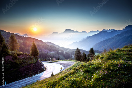Scenic road curving through an alpine landscape with sun shining over the mountains in the Passo Valles area, Dolomites, Italy