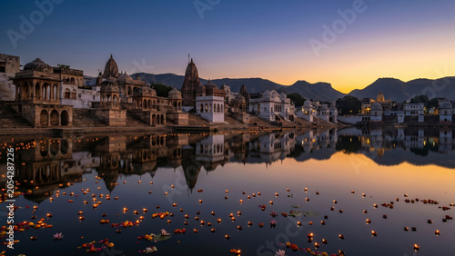 Sacred Pushkar lake ghats at pre-dawn with tiered stone pavilions reflecting in still water saffron gold horizon floating diya offerings indigo sky and lotus on perfectly still surface