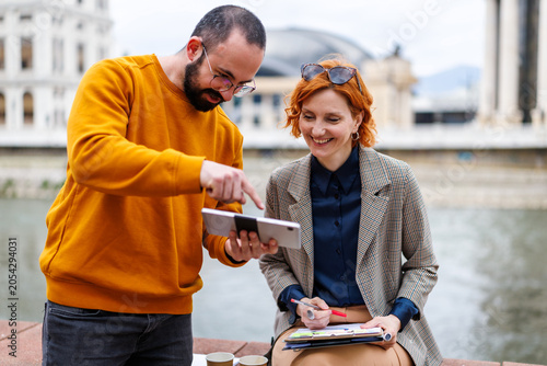 Two people are working together by the river while looking at a tablet in a city during the day
