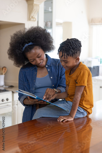 African American mother and son sitting at polished wooden kitchen counter reading picture book