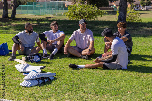 Photos Diverse male teammates sitting on grass at sports field with cricket pads, helme