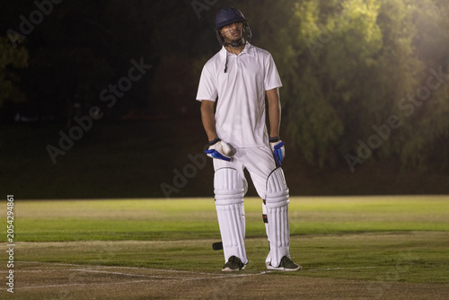 Cricket helmet and bat are resting on floodlit grassy pitch at night near crease lines