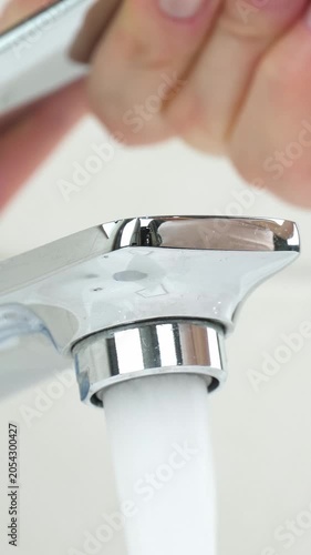 Close-up of water flowing from modern faucet. Hand turns handle to adjust water flow. Clean and fresh water concept in kitchen or bathroom. Vertical angle captures details.