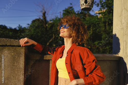 Foto Curly haired woman wearing a red jacket and yellow crop top leans on a stone rai