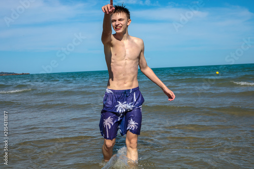 Fotografija Smiling teenage boy standing in sea shallows with one arm raised and finger poin