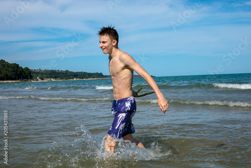 Принти на полотні Smiling teenage boy running through sea shallows holding seaweed, side view, coa
