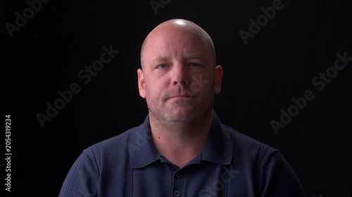 Middle-aged white bald man wearing a dark blue collared shirt with a plain black background, expressionless. 